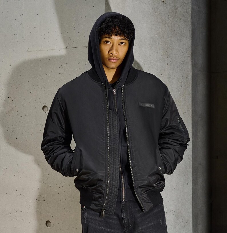 Puede incluir: Chaqueta bomber negra con cierre de cremallera, bolsillos laterales y un peque&ntilde;o parche rectangular en el pecho. La chaqueta se lleva sobre una sudadera con capucha negra y vaqueros negros. El fondo es una pared gris texturizada.