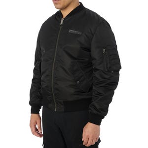 Puede incluir: Chaqueta bomber negra con cremallera, cuello, pu&ntilde;os y dobladillo de canal&eacute;. Cuenta con un peque&ntilde;o bolsillo en la manga izquierda y un parche rectangular sobre el pecho. La chaqueta est&aacute; hecha de un material liso y brillante.
