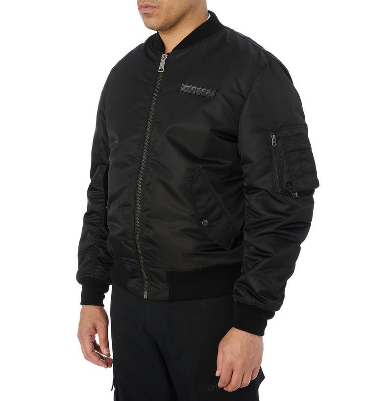 Puede incluir: Chaqueta bomber negra con cremallera, cuello, pu&ntilde;os y dobladillo de canal&eacute;. Cuenta con un peque&ntilde;o bolsillo en la manga izquierda y un parche rectangular sobre el pecho. La chaqueta est&aacute; hecha de un material liso y brillante.