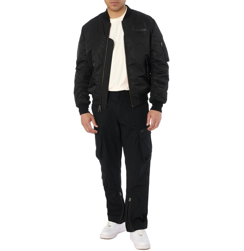 Puede incluir: Una chaqueta bomber negra con cierre de cremallera, usada sobre una camiseta crema. El conjunto incluye pantalones cargo negros con m&uacute;ltiples bolsillos y cremalleras en los tobillos. Zapatillas blancas completan el look.
