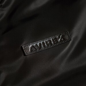 Puede incluir: Primer plano de una chaqueta negra con un parche de cuero. El parche presenta la palabra "AVIREX" en relieve. La tela de la chaqueta tiene un brillo sutil, y la est&eacute;tica general es elegante y minimalista.