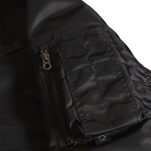 Puede incluir: Primer plano de una chaqueta bomber negra con un bolsillo con cremallera y una cremallera met&aacute;lica. La chaqueta tiene una textura suave y brillante. El bolsillo es rectangular con detalles cosidos. El dise&ntilde;o general es elegante y moderno.
