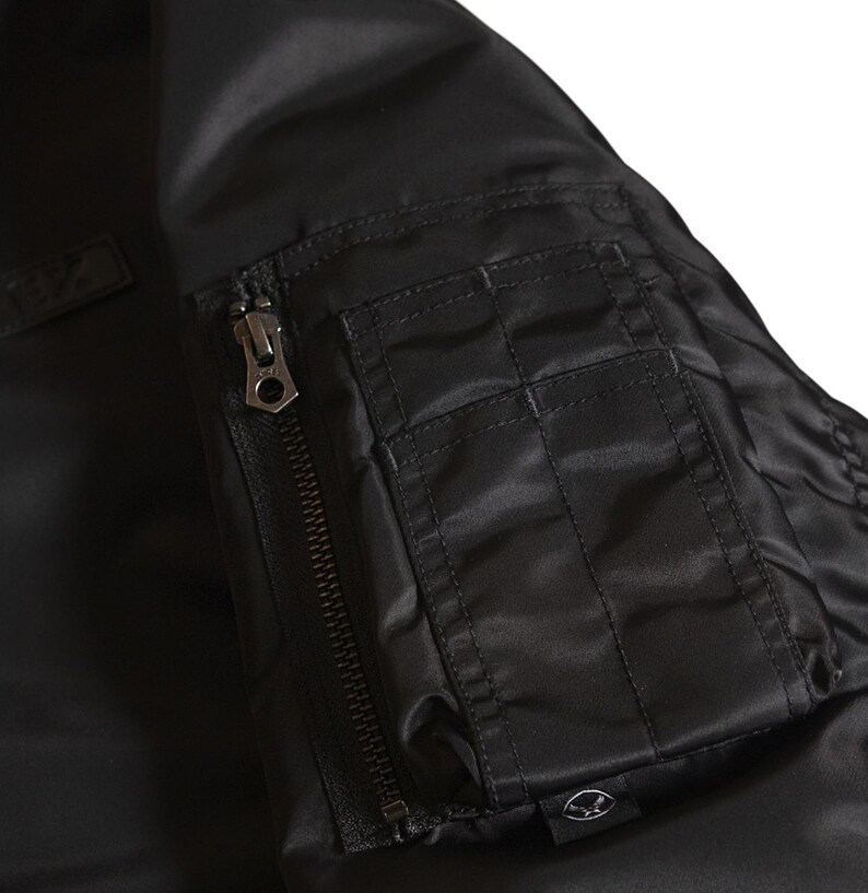 Puede incluir: Primer plano de una chaqueta bomber negra con un bolsillo con cremallera y una cremallera met&aacute;lica. La chaqueta tiene una textura suave y brillante. El bolsillo es rectangular con detalles cosidos. El dise&ntilde;o general es elegante y moderno.