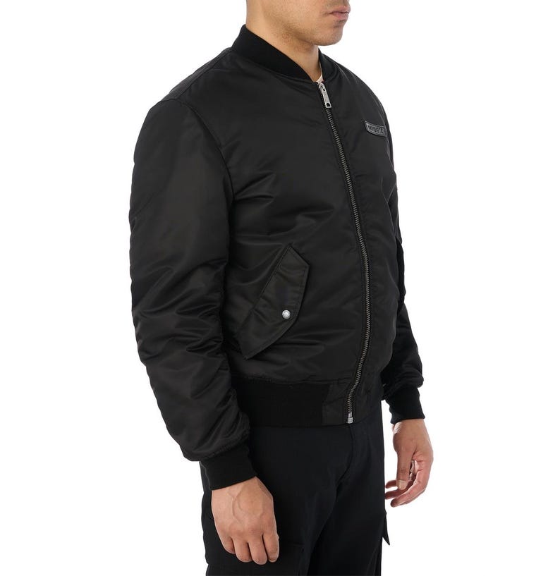 Puede incluir: Chaqueta bomber negra con cremallera, pu&ntilde;os de canal&eacute; y un peque&ntilde;o bolsillo en la manga izquierda. La chaqueta tiene un peque&ntilde;o parche en el pecho. La chaqueta est&aacute; hecha de un material liso y brillante.
