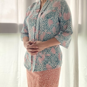 Puede incluir: Conjunto de blusa y falda estampadas. La blusa presenta un diseño geométrico con detalles en rosa y verde azulado sobre fondo blanco. La falda es de color rojo anaranjado con un estampado de hojas. El conjunto lo lleva una persona.