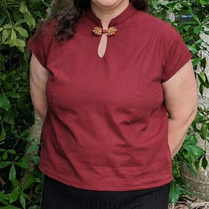 Puede incluir: Una blusa de color rojo oscuro de manga corta con cuello mao y escote tipo cerradura. La prenda se cierra en el cuello con un broche decorativo de metal dorado con forma de dos hojas simétricas situado sobre la abertura del escote.