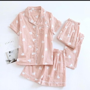 Puede incluir: Un conjunto de pijama rosa claro con estampados de corazones blancos. El conjunto incluye una camisa abotonada de manga corta, un pantalón corto y un pantalón largo. El pijama es de material suave y tiene ribetes blancos.