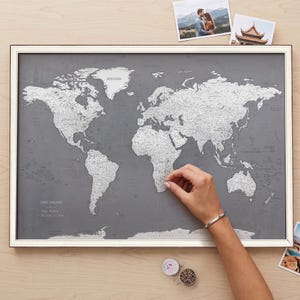 Puede incluir: Un mapa del mundo enmarcado con un fondo gris oscuro y masas terrestres de color plateado. El mapa presenta el texto "QUIET PASSAGES" en la esquina inferior izquierda, junto con una leyenda que enumera "Paths Walked" y "Destinations to Seek".