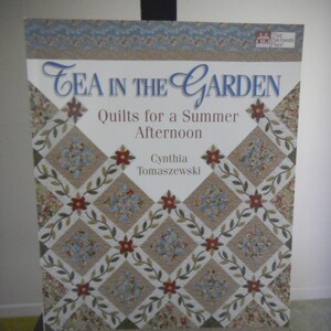 Puede incluir: Portada de libro con el título "Tea in the Garden: Quilts for a Summer Afternoon" de Cynthia Tomaszewski. La portada presenta un diseño de colcha con un patrón floral en tonos rojos, blancos y marrones.
