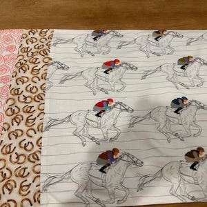 May include: Fabric with three distinct patterns. One features pink roses, another has brown horseshoes, and the third displays racing horses with jockeys in various coloured silks. The horse racing pattern has a white background with grey stripes.