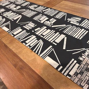 May include: A rectangular table runner with a black background and a repeating white geometric pattern. The pattern includes lines, rectangles, and V-shapes. The runner is placed on a wooden table.