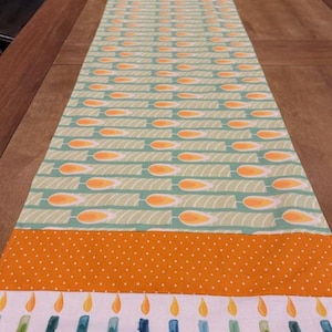 May include: A rectangular table runner with a festive design. The runner features a repeating pattern of orange and yellow candles on a light green background. An orange polka dot section and a white border with candles complete the design.