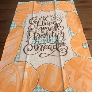 Puede incluir: Un paño de cocina rectangular con un borde a cuadros azul claro y blanco. Presenta ilustraciones de varios panes en tonos naranja. El centro del paño tiene el texto "I love the smell of freshly baked bread" en una fuente decorativa.