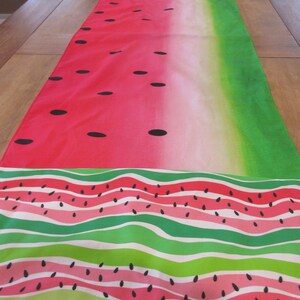 Può includere: Un runner da tavolo a tema anguria con un design sfumato rosso e verde e semi di anguria neri. Il runner ha un motivo ripetitivo di strisce verdi e bianche con fette di anguria rosa e semi neri.