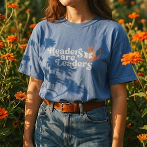 May include: A light blue short-sleeve t-shirt featuring the white text "Readers are Leaders" and a graphic of an orange cat holding an open book. The t-shirt is paired with blue denim jeans and a brown leather belt with a metal buckle.