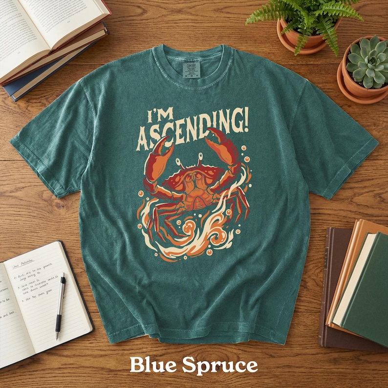 Peut inclure: T-shirt bleu &eacute;pic&eacute;a avec un motif de crabe et le texte "I'M ASCENDING!". Le crabe est orange et rouge, entour&eacute; de flammes orange et blanches. Le t-shirt est sur une surface en bois avec des livres et des plantes.