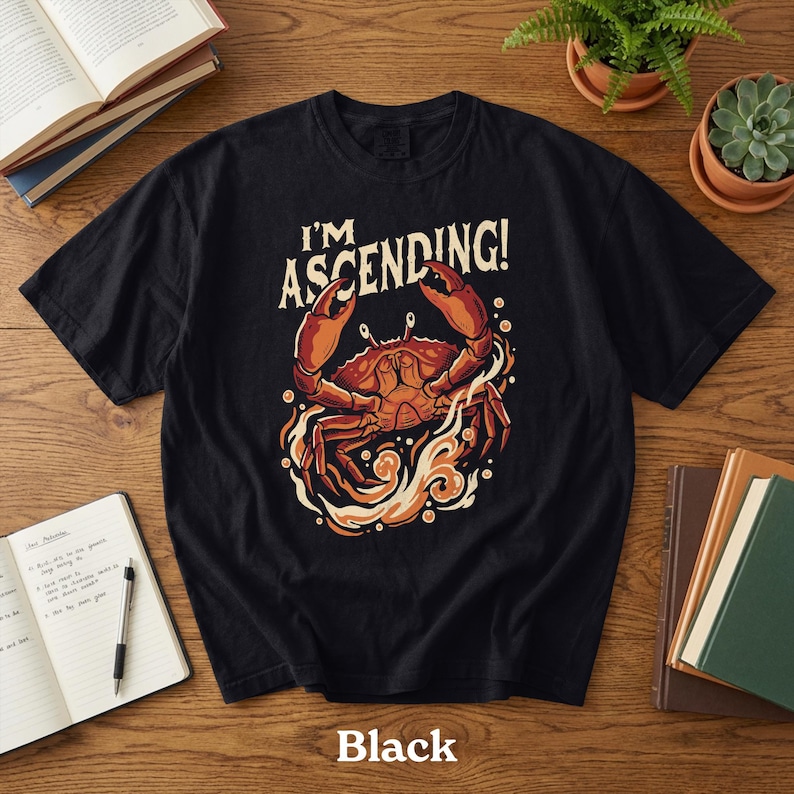 Peut inclure: T-shirt noir avec un motif de crabe et le texte "I'M ASCENDING!". Le crabe est repr&eacute;sent&eacute; en orange et rouge, entour&eacute; de flammes stylis&eacute;es. Le mot "Black" est imprim&eacute; en bas du t-shirt.
