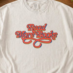 Puede incluir: Sudadera de cuello redondo de color beige claro con la frase "Read More Books" en una fuente retro. El texto es rojo con contornos amarillos y azules. La sudadera tiene una etiqueta en el cuello.