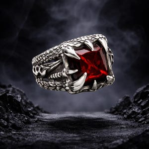 Puede incluir: Anillo plateado con una gran gema roja cuadrada sujeta por garras. El anillo tiene una banda texturizada y detallada con elementos decorativos. El diseño evoca una estética fantástica o gótica, con un aspecto llamativo.