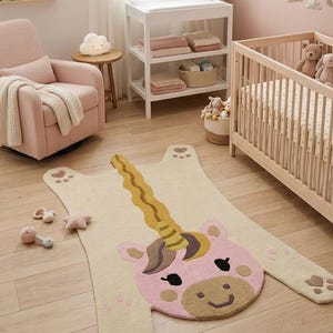 Könnte beinhalten: Cremefarbener Einhorn-Teppich mit rosa Gesicht, braun-gelbem Horn und pfotenförmigen Füßen. Der Teppich befindet sich in einem Kinderzimmer mit einem Kinderbett, einer Wickelkommode und einem Schaukelstuhl. Einige Babyspielzeuge sind verstreut.
