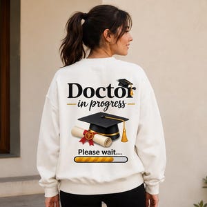 Peut inclure: Un sweat-shirt blanc à col rond avec un motif au dos portant l'inscription Doctor in progress, une toque de diplômé noire, un diplôme enroulé avec un sceau rouge et une barre de chargement avec le texte Please wait sous le motif.