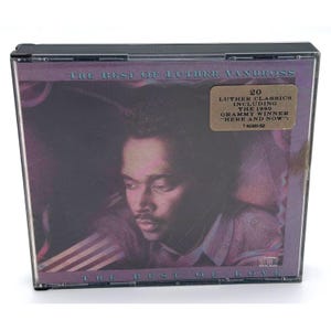 Puede incluir: Una caja de CD con el álbum "The Best of Luther Vandross". La portada presenta un retrato de Luther Vandross en un diseño en tonos morados. Una pegatina indica que el álbum incluye la canción ganadora del Grammy de 1990 "Here and Now".