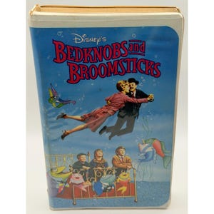 May include: A Disney Bedknobs and Broomsticks VHS tape with a blue cover featuring a man and woman flying through the air above three children sitting on a bed surrounded by colorful cartoon fish and bubbles against a light blue background.