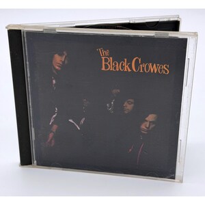 May include: A clear plastic CD jewel case containing The Black Crowes music album. The cover art features a dark, moody photograph of band members in black clothing against a black background, with the band name printed in orange text.