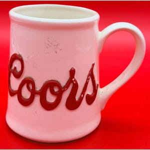 Puede incluir: Una taza de cerámica rosa vintage con interior blanco y asa curva. La palabra "Coors" está escrita en letras rojas en relieve en la parte delantera de la taza. La taza está sobre un fondo rojo.
