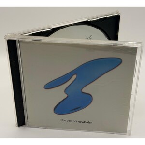 Puede incluir: Una caja de CD de plástico transparente que contiene el álbum (the best of) NewOrder. La portada presenta un fondo gris claro con una gran forma orgánica azul abstracta en el centro. El disco es parcialmente visible dentro de la caja abierta.