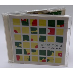Pode incluir: Uma caixa de CD de plástico transparente contendo o álbum A better version of me de Rainer Maria. A capa apresenta um padrão de grade com formas amarelas, verde-escuras e vermelhas sobre um fundo branco com o texto impresso.