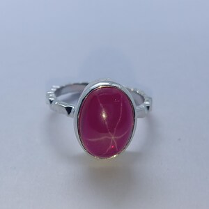 May include: A silver ring featuring a polished oval-shaped hot pink cabochon gemstone. The stone displays a distinct six-rayed star pattern across its surface. The metal band has a textured, notched edge design surrounding the central setting.