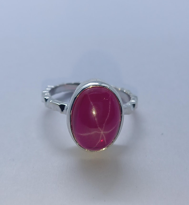 May include: A silver ring featuring a polished oval-shaped hot pink cabochon gemstone. The stone displays a distinct six-rayed star pattern across its surface. The metal band has a textured, notched edge design surrounding the central setting.