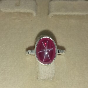 May include: A silver ring featuring a polished, oval-shaped, deep magenta star gemstone. The stone displays a distinct six-rayed white star pattern across its smooth, domed surface. The metal band is simple and polished, holding the stone in a bezel setting.