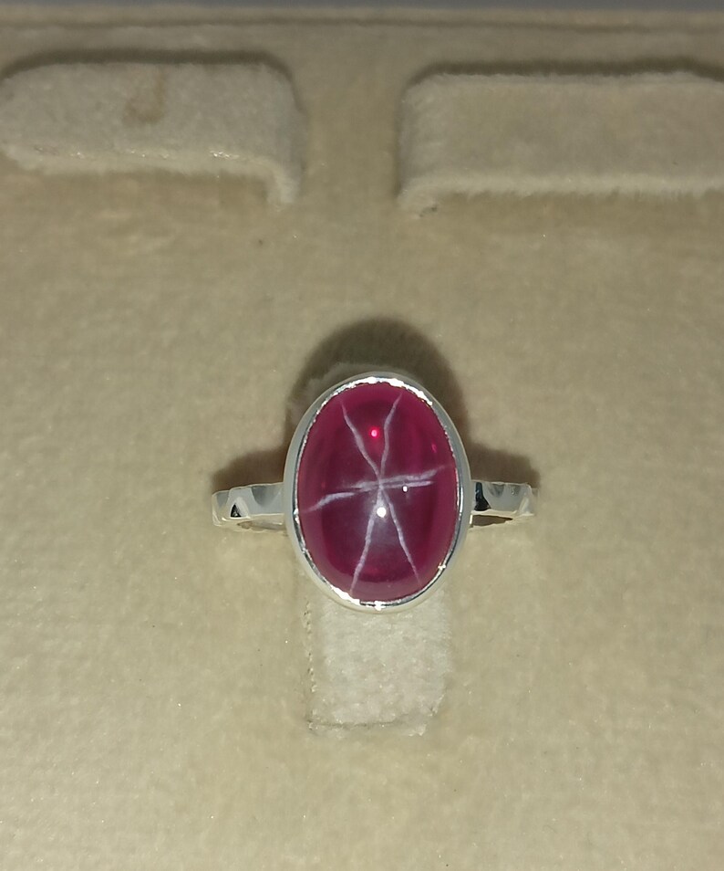 May include: A silver ring featuring a polished, oval-shaped, deep magenta star gemstone. The stone displays a distinct six-rayed white star pattern across its smooth, domed surface. The metal band is simple and polished, holding the stone in a bezel setting.