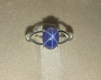 May include: A silver-toned ring featuring a central oval-shaped blue cabochon gemstone with a distinct six-rayed star pattern. The metal band has a split design on both sides of the stone, secured by four prongs holding the gem in place.