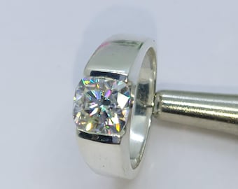 May include: A silver-toned ring featuring a single, square-cut, clear gemstone with multifaceted edges that reflect light in a spectrum of colours. The band is smooth, polished, and wide, securely holding the central stone in a flush setting.