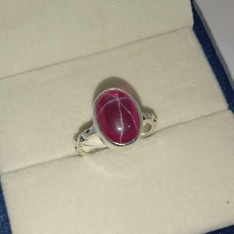 May include: A silver ring featuring an oval-shaped, deep magenta star gemstone set in a smooth bezel. The band of the ring has a decorative, textured pattern along the sides, providing a detailed contrast to the polished metal and the stone.