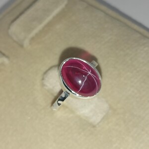 May include: A silver ring featuring a polished, oval-shaped deep pink gemstone with a visible six-rayed star effect across its surface. The stone is set in a smooth, plain silver bezel mount, and the band is made of a simple, polished silver metal.