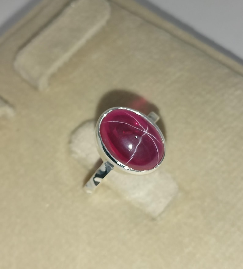 May include: A silver ring featuring a polished, oval-shaped deep pink gemstone with a visible six-rayed star effect across its surface. The stone is set in a smooth, plain silver bezel mount, and the band is made of a simple, polished silver metal.