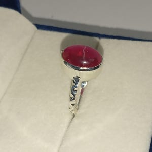 May include: A silver ring featuring a smooth, oval-shaped, deep red cabochon gemstone with a thin white line running across its surface. The metal band has a decorative, cut-out geometric pattern along the sides, providing a detailed texture.