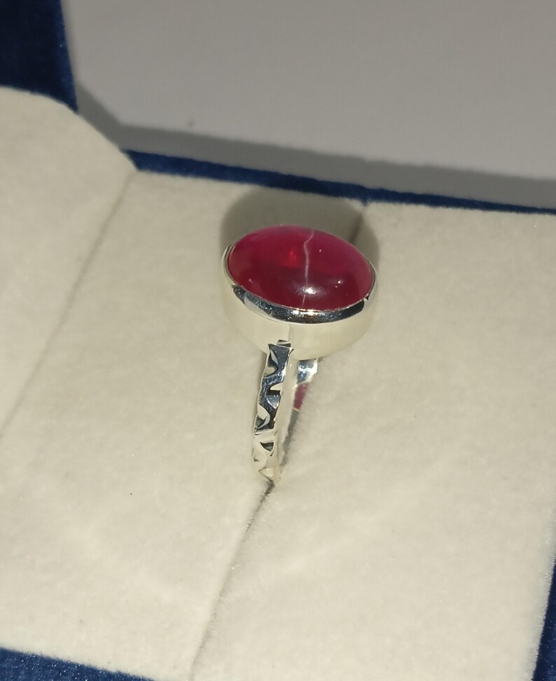 May include: A silver ring featuring a smooth, oval-shaped, deep red cabochon gemstone with a thin white line running across its surface. The metal band has a decorative, cut-out geometric pattern along the sides, providing a detailed texture.