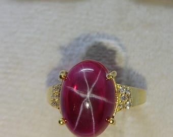 May include: A gold ring featuring a large, oval-shaped, deep pink star ruby cabochon with a visible six-rayed star effect. The central gemstone is held by four prongs, and the band is accented with small, clear, round-cut stones on both sides.
