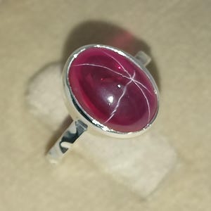 May include: A silver ring featuring a polished, oval-shaped deep pink star gemstone set in a smooth metal bezel. The stone displays a distinct six-rayed star pattern across its surface, held by a simple, slender, polished silver band.
