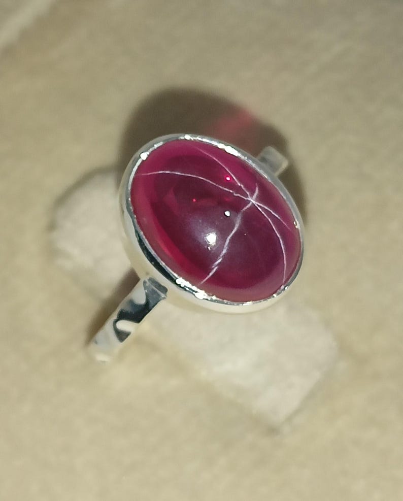 May include: A silver ring featuring a polished, oval-shaped deep pink star gemstone set in a smooth metal bezel. The stone displays a distinct six-rayed star pattern across its surface, held by a simple, slender, polished silver band.