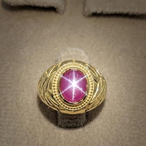 Könnte beinhalten: Ein goldener Ring mit einem ovalen Sternrubin-Cabochon in der Mitte. Der Stein zeigt einen ausgeprägten sechsstrahligen Sterneffekt. Die Ringschiene ist mit einem strukturierten, gravierten Muster und einem Perlenrand um den Stein versehen.
