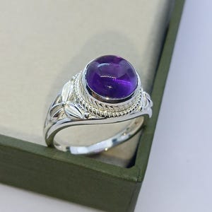 May include: A silver ring featuring a smooth, round, purple amethyst cabochon stone set in a decorative bezel. The band is crafted with a twisted rope texture surrounding the stone and detailed leaf-like engravings along the sides of the shank.