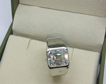 May include: A silver-toned ring featuring a large, rectangular emerald-cut clear gemstone set in a smooth, polished metal band. The ring is positioned upright, showcasing the faceted stone's reflective surface and the solid, wide metal shank design.