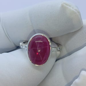 May include: A silver ring featuring a large, oval-shaped, deep pink star gemstone set in a smooth metal bezel. The band of the ring has a textured, hammered-style finish along the sides, providing a contrast to the polished setting of the stone.
