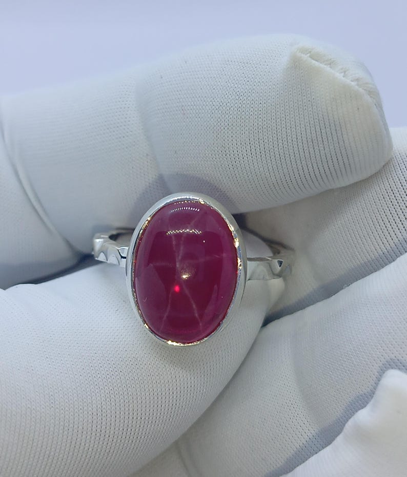 May include: A silver ring featuring a large, oval-shaped, deep pink star gemstone set in a smooth metal bezel. The band of the ring has a textured, hammered-style finish along the sides, providing a contrast to the polished setting of the stone.